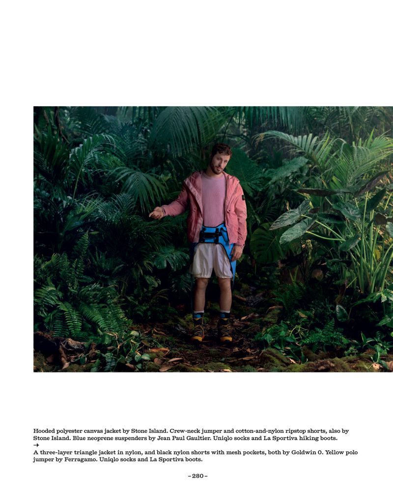 Man standing in lush jungle foliage wearing a pink jacket, white shorts, blue suspenders, and hiking boots.