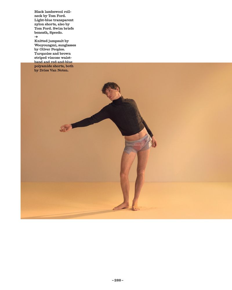 Model posing barefoot in a black roll-neck top and sheer light-blue shorts against a warm tan studio backdrop.