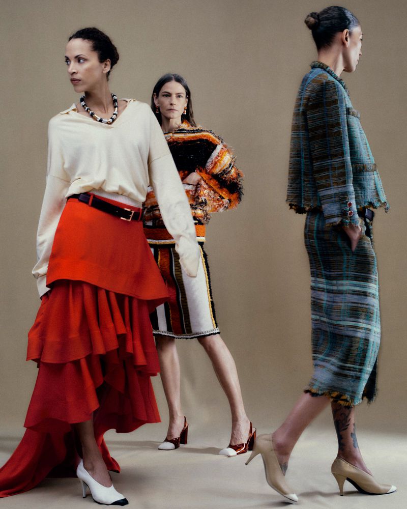 Three models wearing high-fashion knitwear, ruffled red skirt, and plaid tweed suits posing against a neutral studio backdrop.