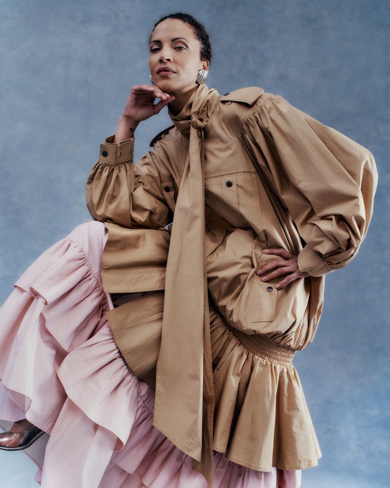Noémie Lenoir posing in a tan balloon-sleeve coat with neck tie and tiered pale pink ruffle skirt against a blue textured backdrop.