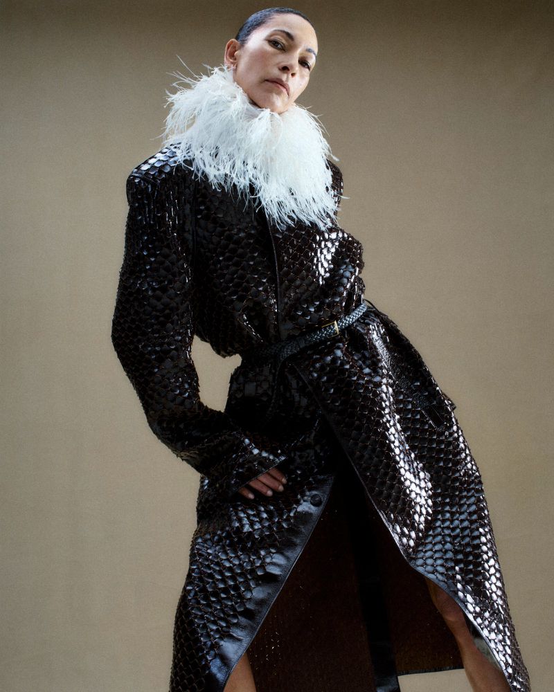 Model Noémie Lenoir wearing a glossy snakeskin coat with a white feather collar against a neutral backdrop.