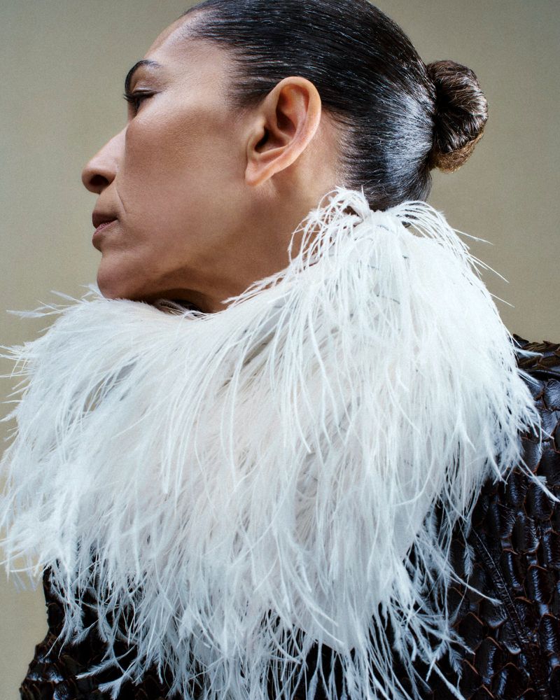 Close-up profile of a woman wearing an oversized white feather collar and dark textured top against a neutral background.