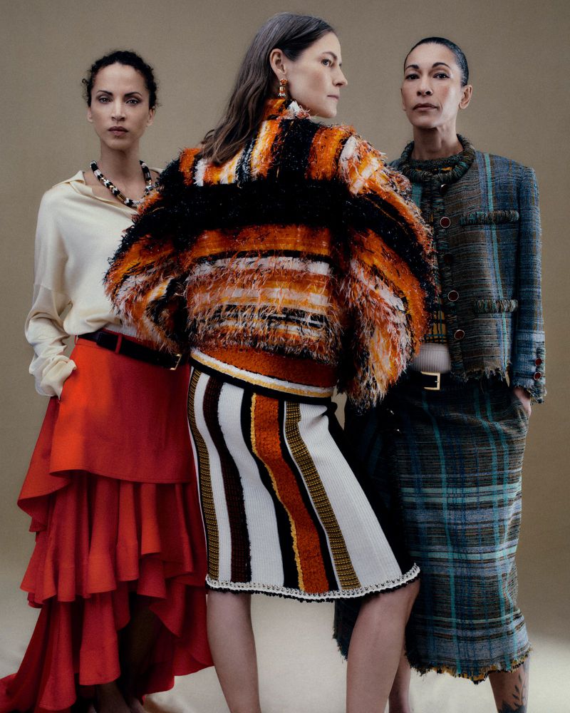 Three models pose in designer knitwear, a red tiered skirt, and a plaid tweed suit against a warm neutral studio backdrop.