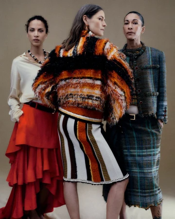Three models pose in designer knitwear, a red tiered skirt, and a plaid tweed suit against a warm neutral studio backdrop.