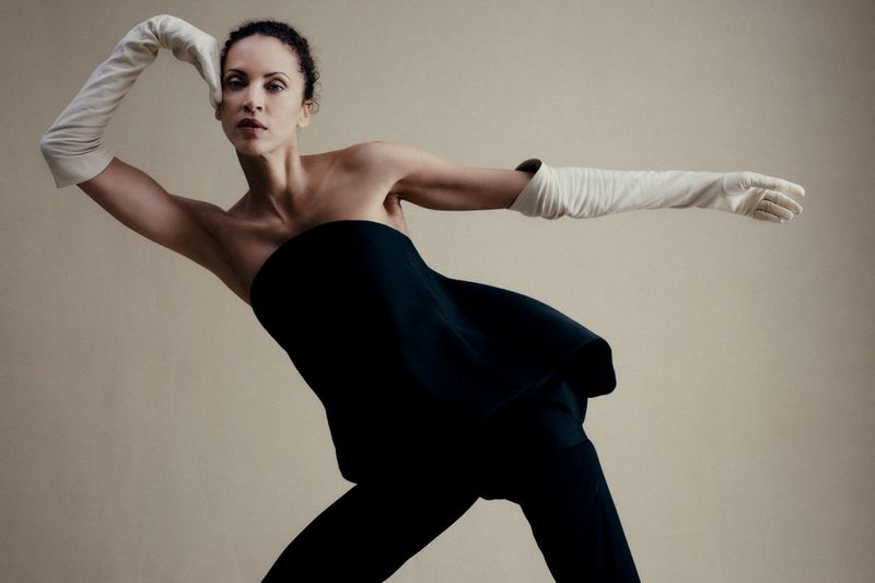 Model Noémie Lenoir poses dynamically in a black strapless top and long cream opera gloves against a neutral studio backdrop.