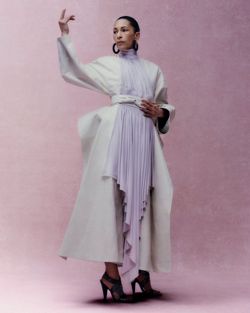 Model posing sculpturally in a structured off-white coat and pleated lilac dress against a mottled pink studio backdrop.