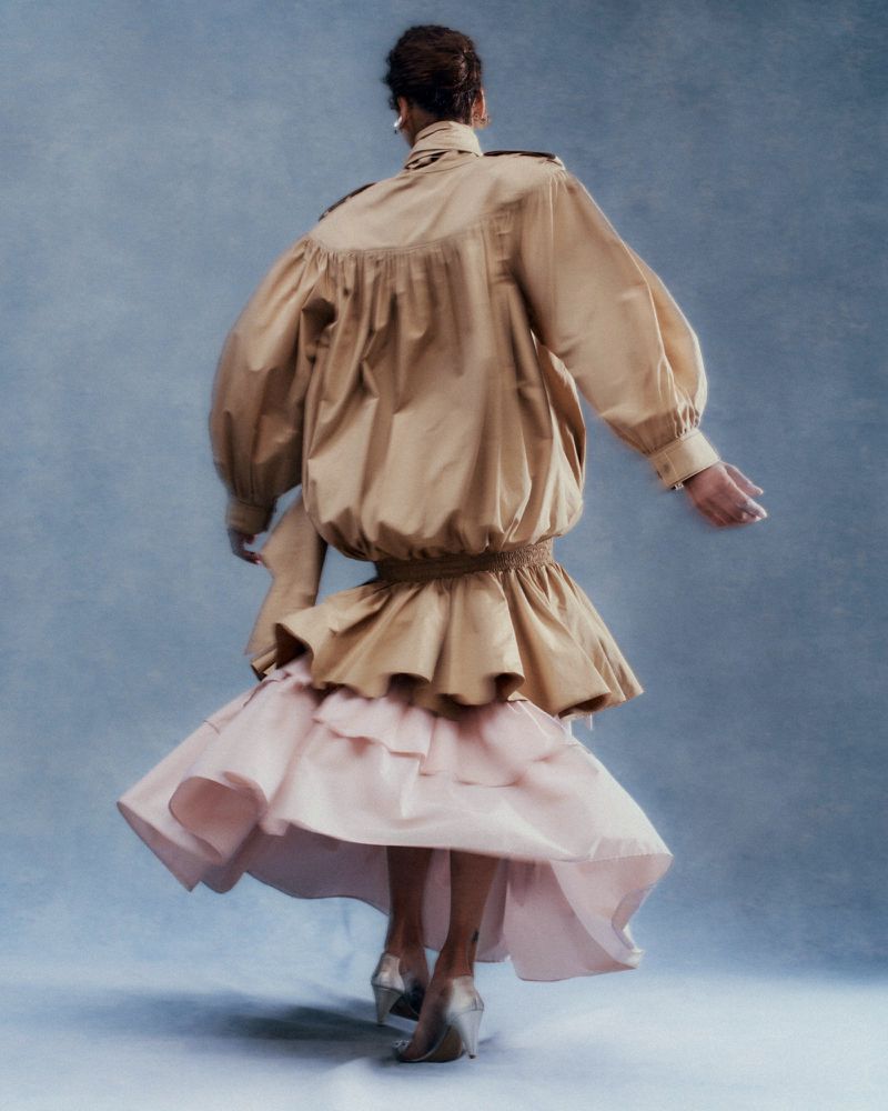 Model seen from behind mid-turn wearing an oversized tan jacket and tiered pale pink ruffled skirt against a blue textured studio backdrop.