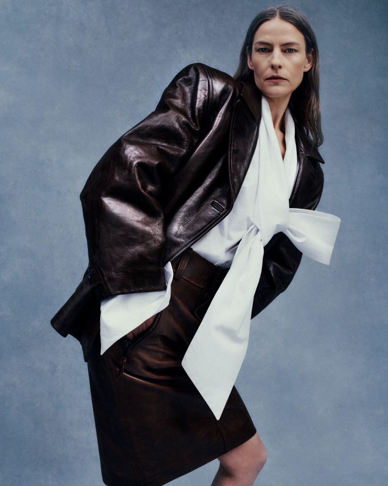 Model in brown leather blazer wearing white blouse with large bow against blue textured studio background