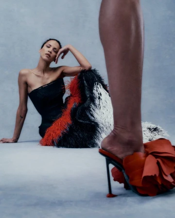 Model in a strapless feather gown seated on a studio floor framed behind a foreground ruffled high-heeled mule.