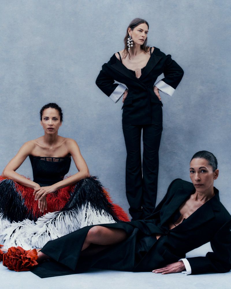 Three models in black tailoring and a feathered gown posing against a textured cool grey studio backdrop.
