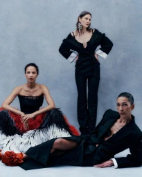 Three models in black tailoring and a feathered gown posing against a textured cool grey studio backdrop.