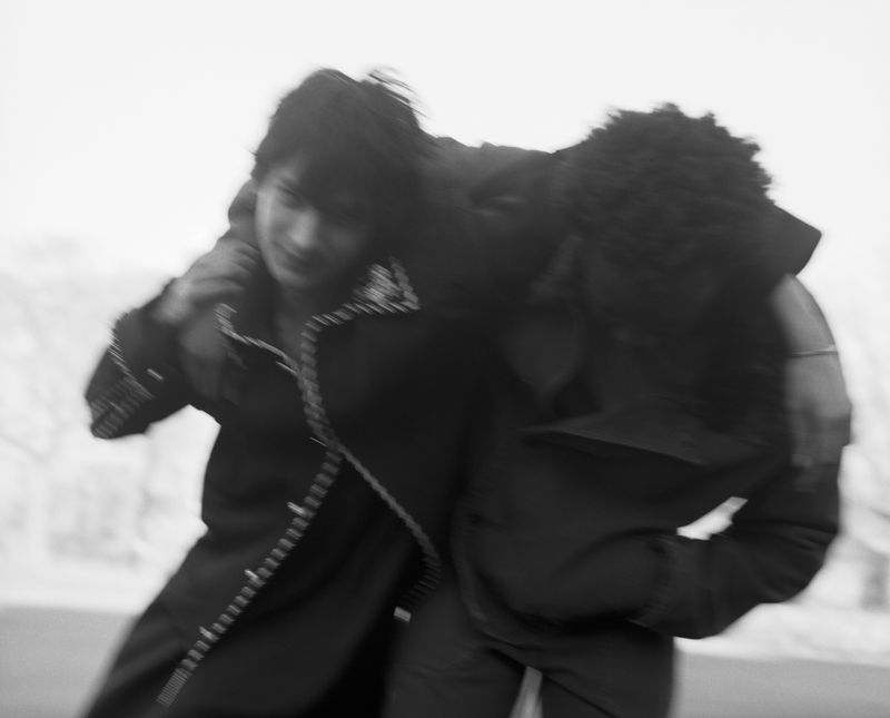 Two men in dark coats leaning on each other in blurred black and white editorial photography.