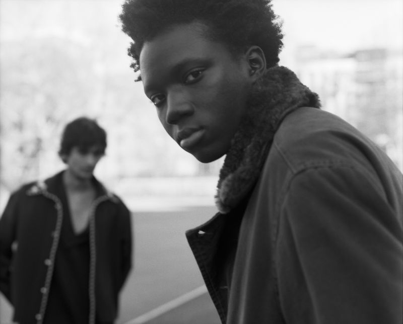 Black and white photo of a young Black man looking over his shoulder outdoors with another man visible in the background.
