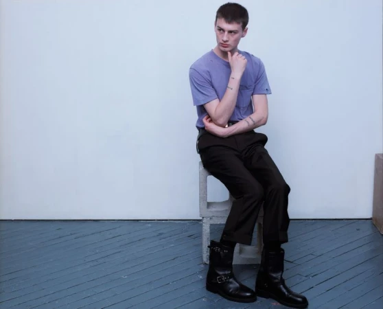 Young man in a purple t-shirt, dark trousers, and black leather boots seated on concrete blocks against a neutral studio wall.