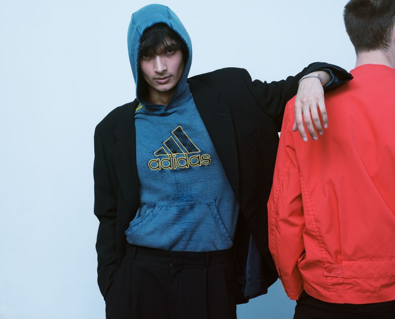 A young man in a blue hoodie under a black blazer rests his arm on another person wearing a red jacket against a pale backdrop.