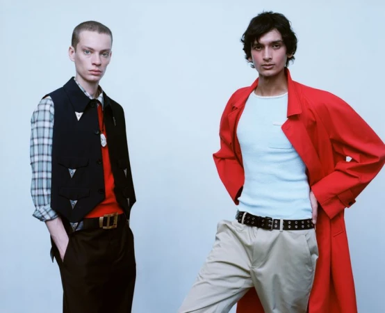 Two male models posing in layered menswear, featuring a black structured vest with silver hardware and a vibrant red coat.