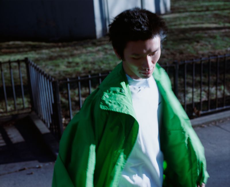An East Asian man walking outside wearing an oversized bright green jacket over a white top.