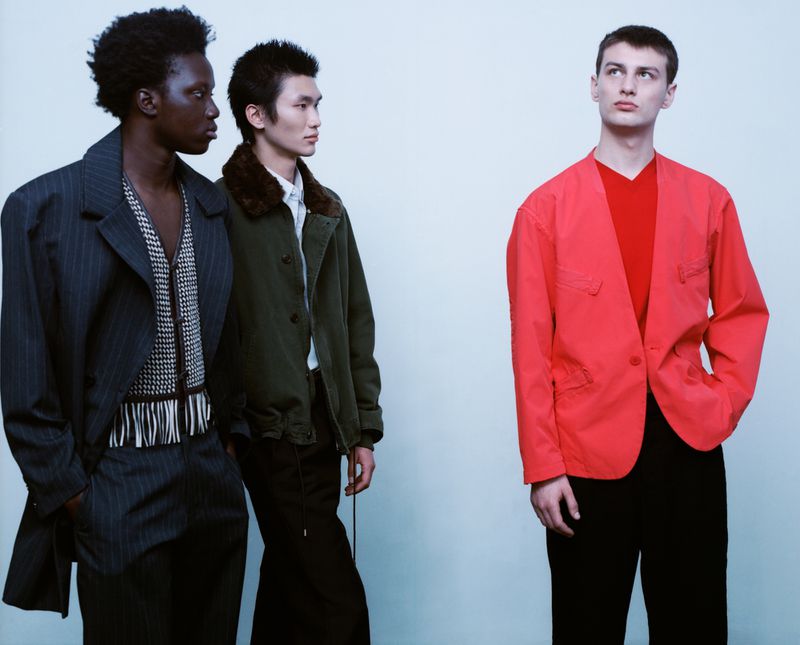 Three male models in contemporary designer menswear including a red jacket pose against a seamless pale studio background.