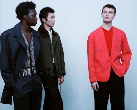 Three male models in contemporary designer menswear including a red jacket pose against a seamless pale studio background.