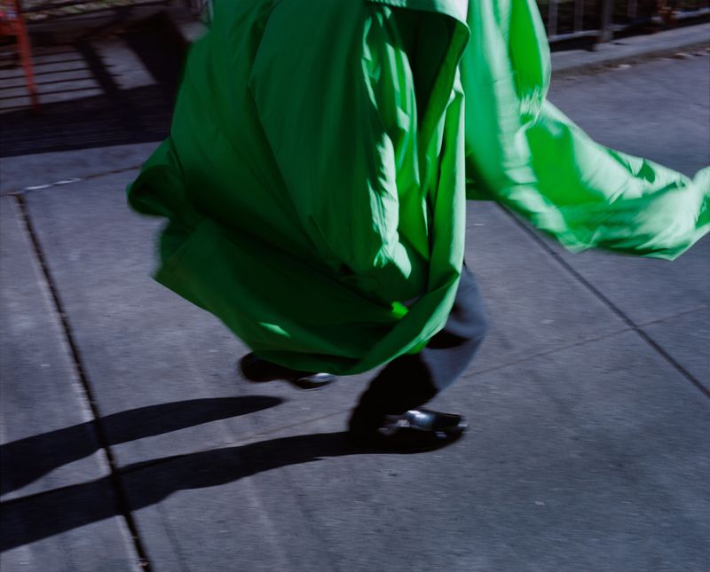 Cropped low-angle view of a person wearing a billowing bright green coat, dark trousers, and black shoes moving across a sidewalk.