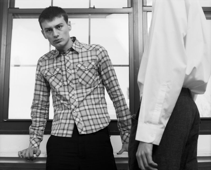 Monochrome fashion editorial image featuring a young man in a plaid shirt leaning against a window with another person in a white shirt