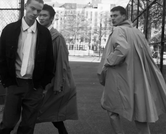 Three male models in tailored outerwear and ties posing outdoors beside a chain-link fence in black and white.