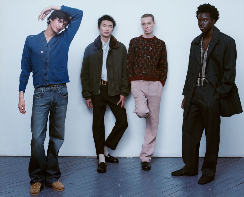 Four male models posing in styled menswear standing in a row against a plain studio wall on painted wooden floorboards.