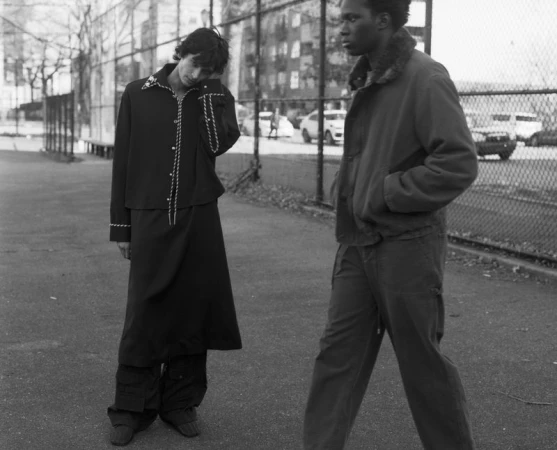 Two male models in dark outerwear stand outdoors beside a chain-link fence in a black-and-white fashion editorial.