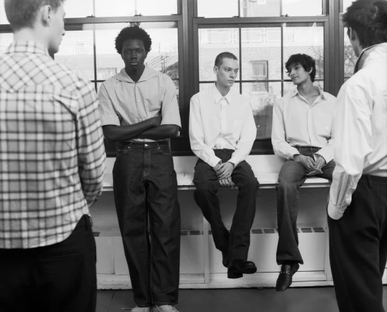Five male models in casual menswear gathered in front of large grid windows in a black and white fashion editorial.