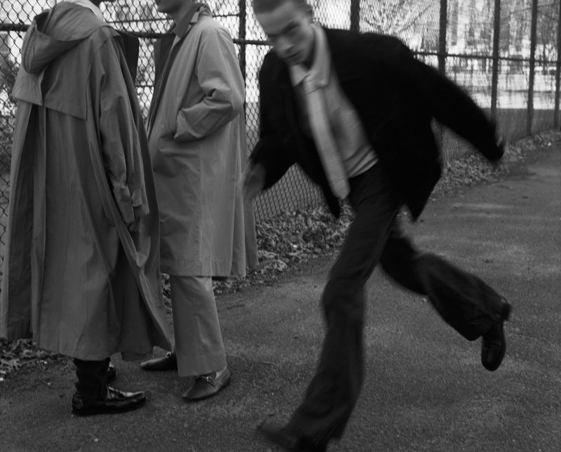Black and white fashion photo of a man in dynamic motion wearing a dark jacket while two figures in long coats stand by a fence.