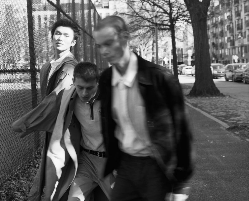 Three young men walking along an urban sidewalk next to a chain-link fence in a black and white fashion editorial photograph.