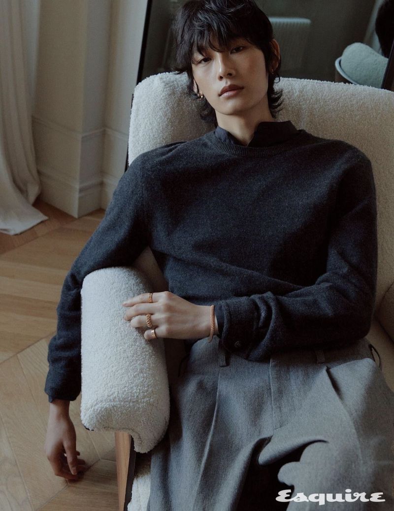 Model Dugyeong Kim wearing a dark grey knit sweater and grey trousers seated in a white boucle armchair.