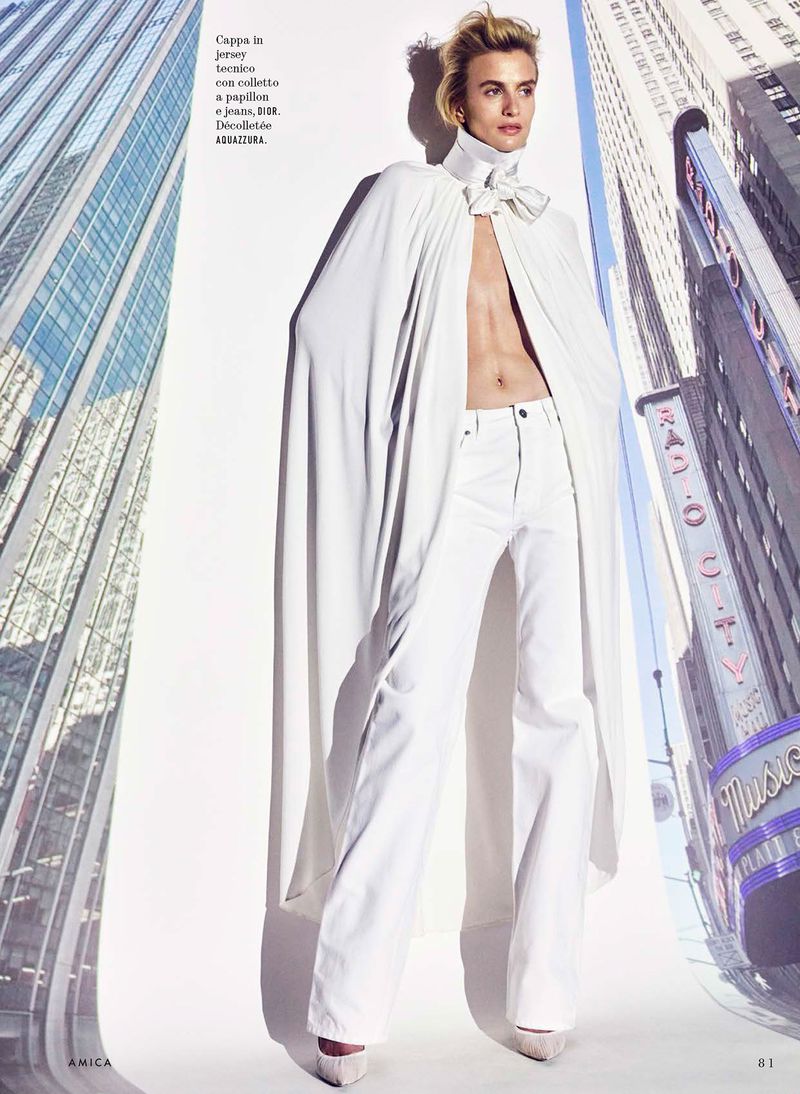 Model Veronika Vilim posing in an open white cape and matching white pants flanked by skyscraper backdrops.