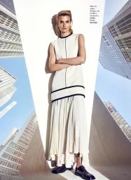 Veronika Vilim in a white sleeveless pleated dress and black derby shoes posing against a skyscraper print backdrop for Amica.