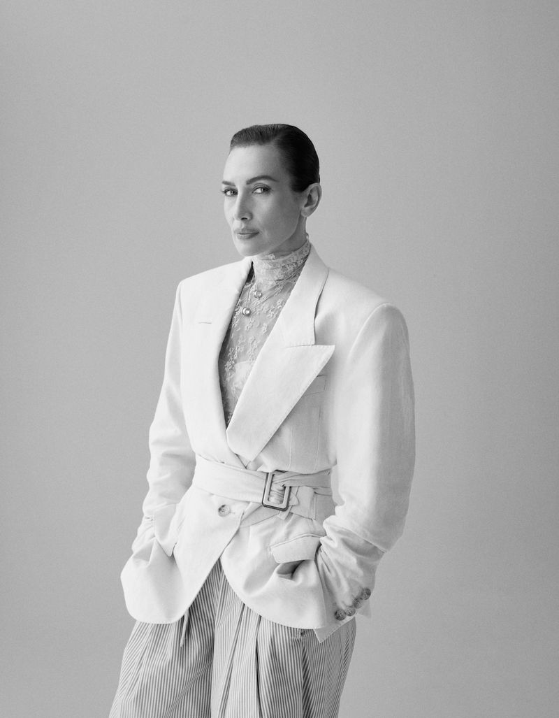 Model Nieves Alvarez posing in a belted white blazer, sheer high-neck top, and pleated trousers against a neutral studio backdrop.
