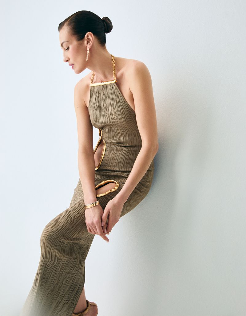 Model Nieves Alvarez wearing an olive textured halter dress with gold cutout details against a pale studio backdrop.