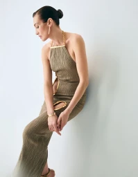 Model Nieves Alvarez wearing an olive textured halter dress with gold cutout details against a pale studio backdrop.