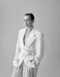 Model Nieves Alvarez posing in a belted white blazer, sheer high-neck top, and pleated trousers against a neutral studio backdrop.