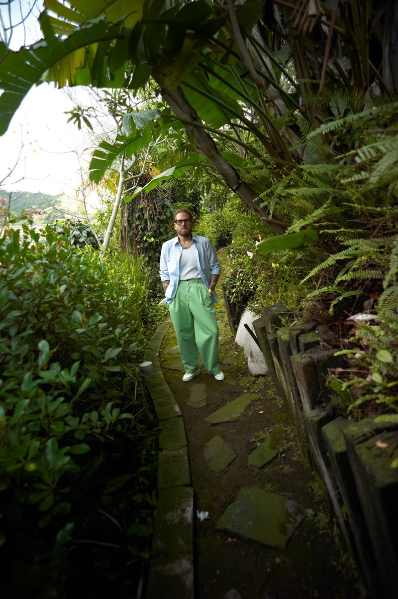 Jonah Hill standing on a lush stone garden path wearing light green trousers and an open light blue button-down shirt.