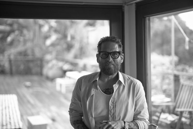 Black and white portrait of Jonah Hill wearing glasses and an open button-down shirt in front of glass doors.