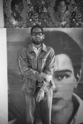 Jonah Hill wearing a denim jacket and dark eyeglasses posing in front of black and white portrait artwork.