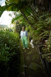 Jonah Hill standing on a lush stone garden path wearing light green trousers and an open light blue button-down shirt.
