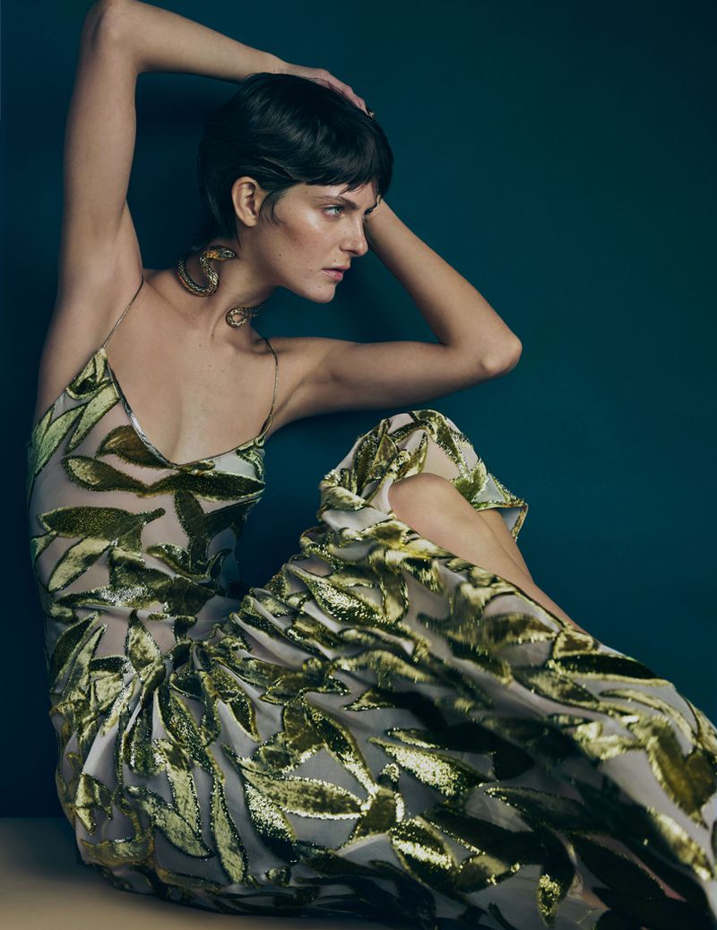 Woman with short dark hair in a sheer green leaf pattern dress and gold snake necklace seated against a deep teal background.