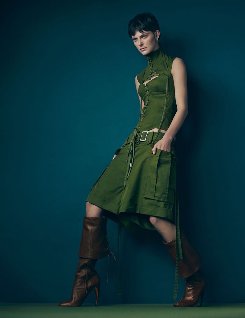 Model in an olive green corset top and matching pleated skirt paired with brown leather heeled boots against a dark teal studio backdrop.