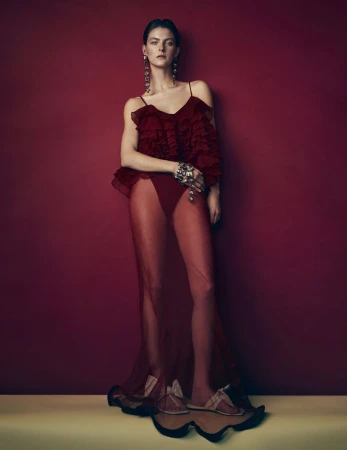 Model standing against a deep burgundy backdrop wearing a sheer red ruffled gown with long statement earrings.