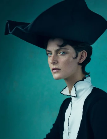 Fashion portrait of a model with short dark hair wearing an oversized black sculptural hat, white collared shirt, and black jacket against teal.