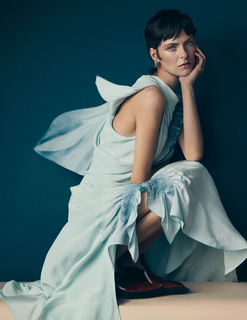 Model with short dark hair crouching in a pale draped gown with ruffles against a dark teal backdrop.