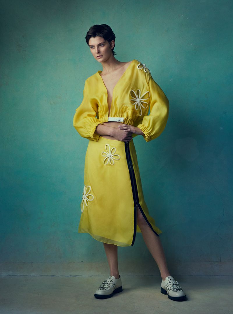 Model with short dark hair wearing a vibrant yellow two-piece ensemble with floral appliques against a textured teal backdrop.