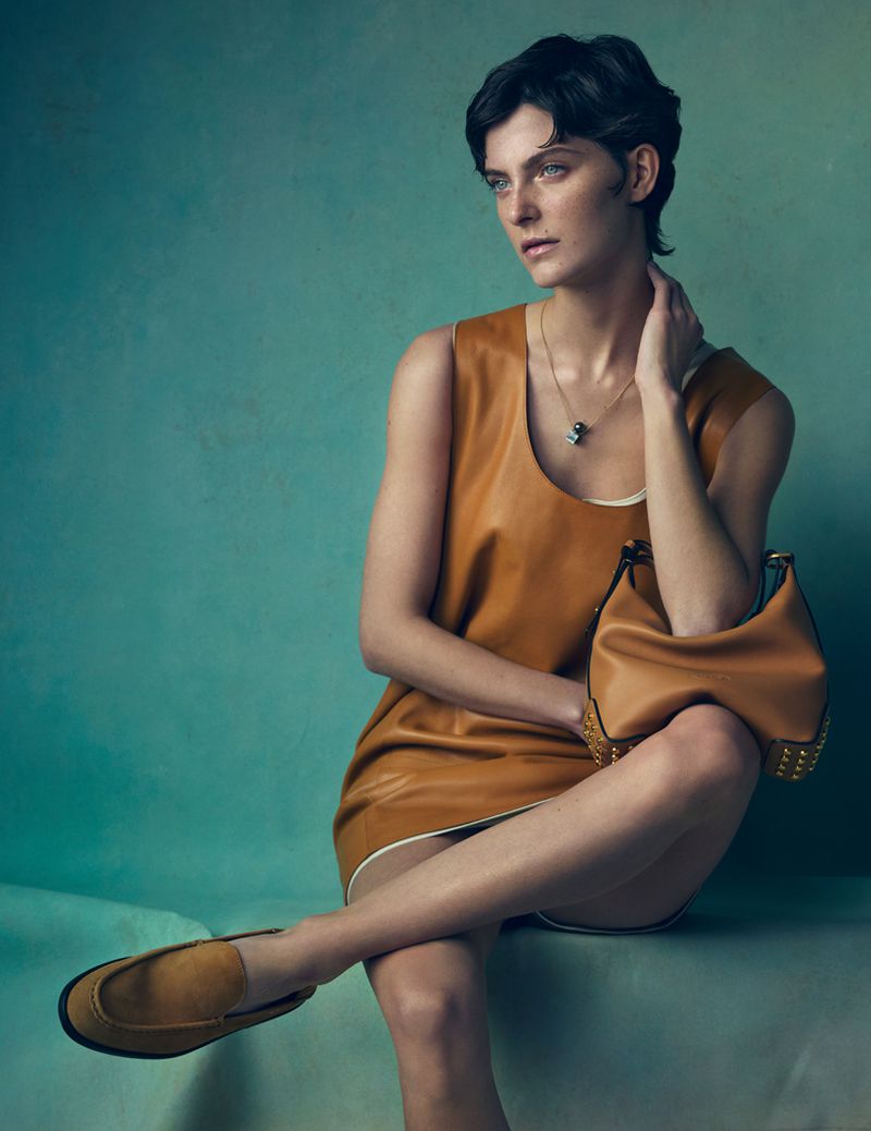 Model with short dark hair wearing a tan leather shift dress and matching loafers seated against a teal textured background.
