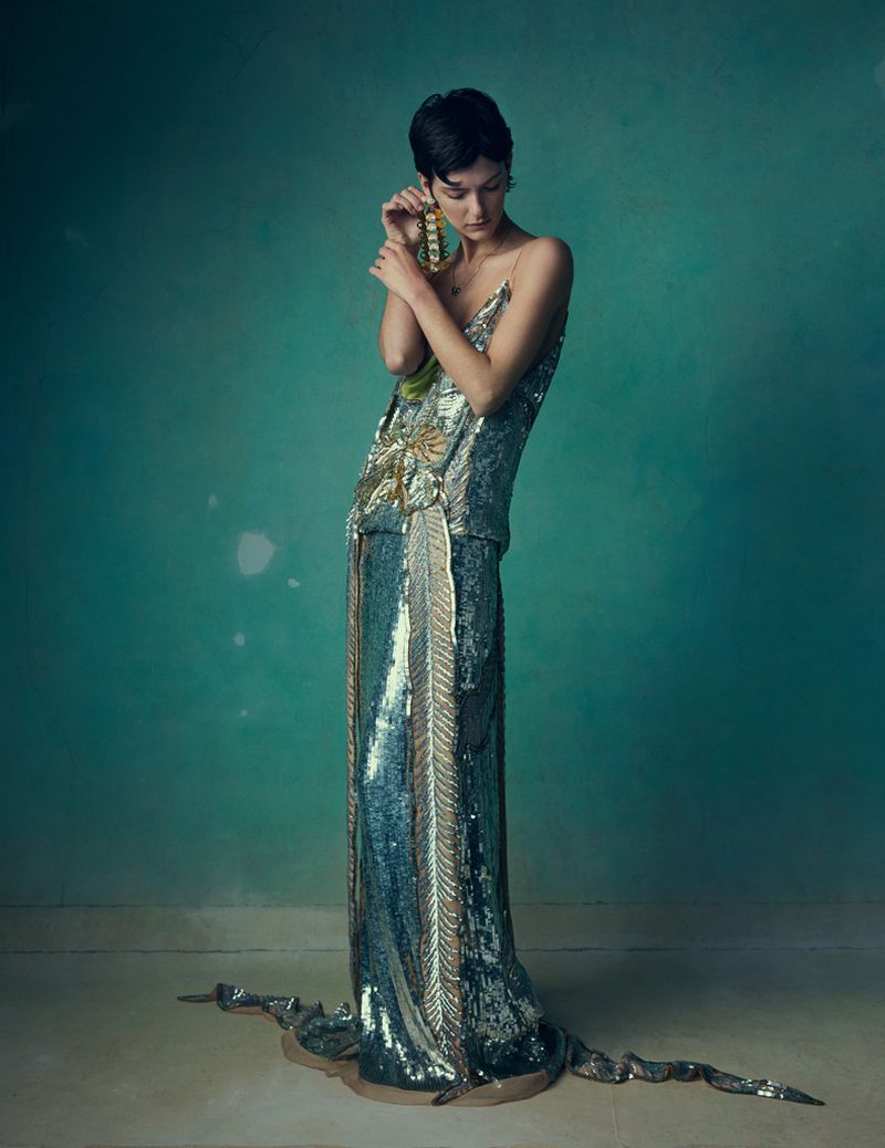 Model with short dark hair wearing an embellished sequined floor-length evening gown against a textured teal backdrop.